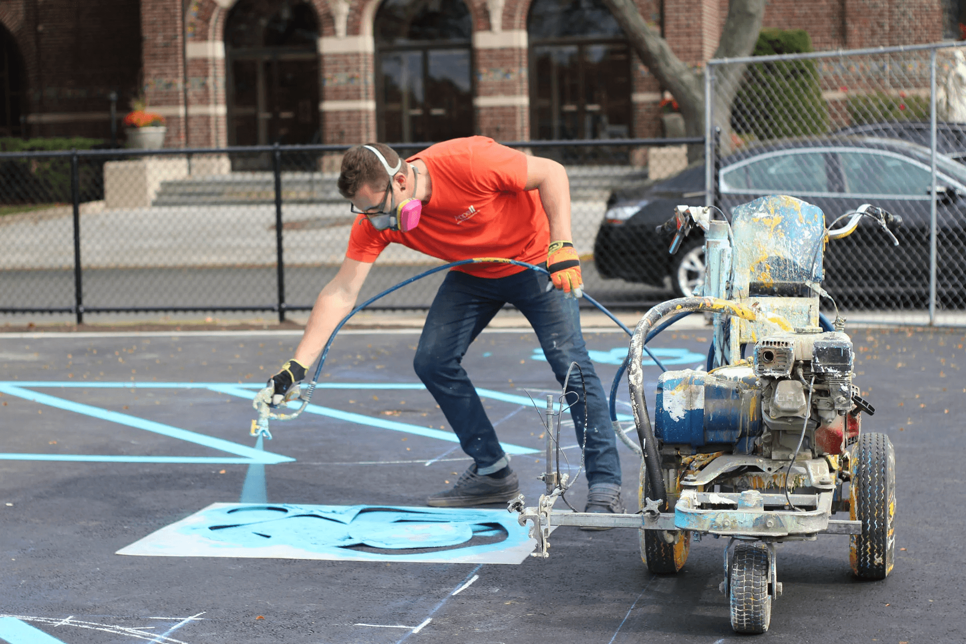 A person using a paint sprayer to apply blue paint to a sports court surface.