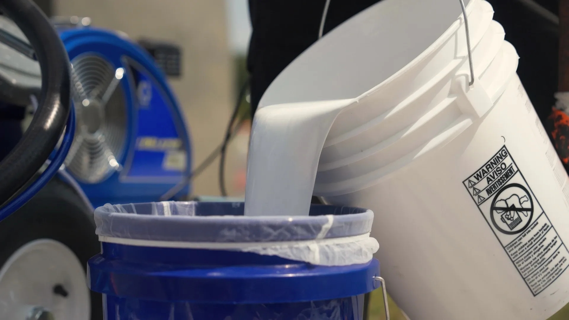 Appell striping vender buying supplies line striping A close-up of white paint being poured into a blue bucket, with a warning label visible on the bucket.
